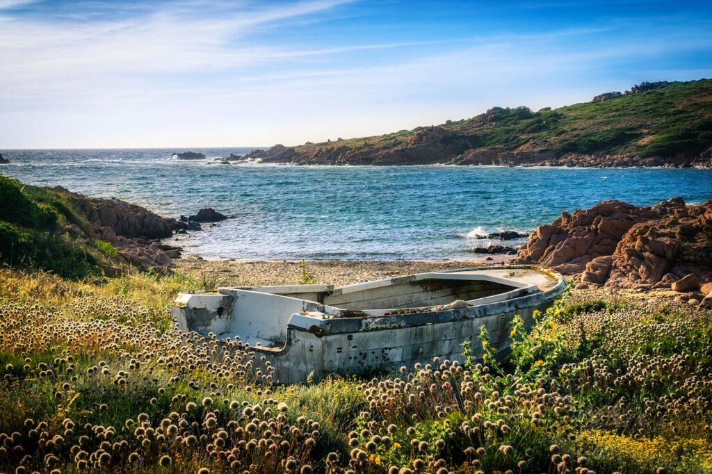 An abandoned boat sits amidst wildflowers on a grassy shore, overlooking the clear blue waters and rocky coastline of Sardinia, one of the Top 5 Italian Islands, under a bright blue sky. This scene captures the island's rugged charm and pristine natural beauty.