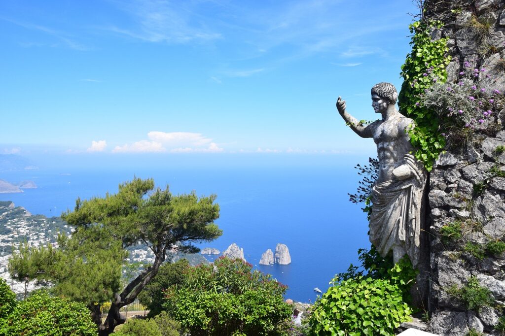 A statue partially covered in ivy overlooks the blue sea and rocky islets of Capri, one of the Top 5 Italian Islands, with a lush green landscape and a clear blue sky in the background. This captivating scene epitomizes the island's natural beauty and serene ambiance.