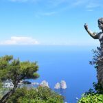 A statue partially covered in ivy overlooks the blue sea and rocky islets of Capri, one of the Top 5 Italian Islands, with a lush green landscape and a clear blue sky in the background. This captivating scene epitomizes the island's natural beauty and serene ambiance.