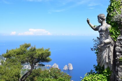 A statue partially covered in ivy overlooks the blue sea and rocky islets of Capri, one of the Top 5 Italian Islands, with a lush green landscape and a clear blue sky in the background. This captivating scene epitomizes the island's natural beauty and serene ambiance.