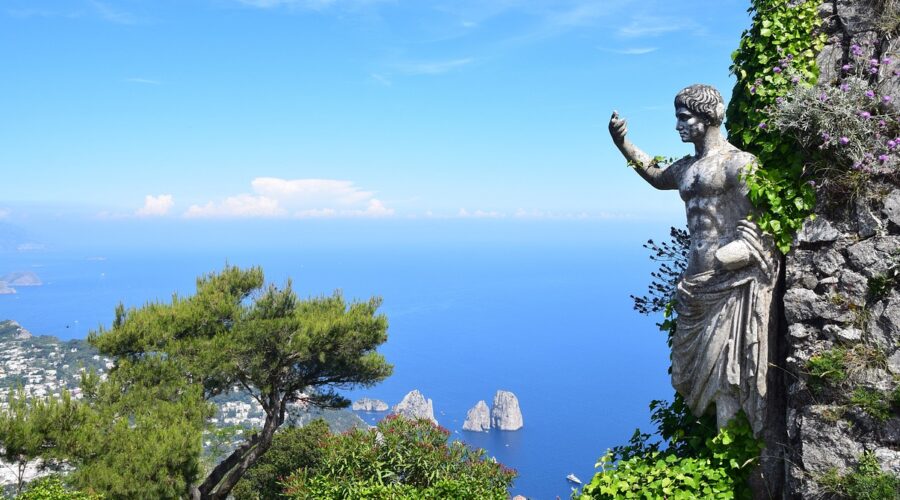 A statue partially covered in ivy overlooks the blue sea and rocky islets of Capri, one of the Top 5 Italian Islands, with a lush green landscape and a clear blue sky in the background. This captivating scene epitomizes the island's natural beauty and serene ambiance.