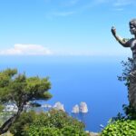 A statue partially covered in ivy overlooks the blue sea and rocky islets of Capri, with a lush green landscape and a clear blue sky in the background.