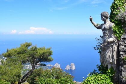 A statue partially covered in ivy overlooks the blue sea and rocky islets of Capri, with a lush green landscape and a clear blue sky in the background.