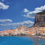 View of Cefalù, Italy, featuring its colorful buildings along the coastline, a prominent cathedral, and a rocky hill under a bright blue sky with fluffy clouds.