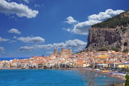 View of Cefalù, Italy, featuring its colorful buildings along the coastline, a prominent cathedral, and a rocky hill under a bright blue sky with fluffy clouds.