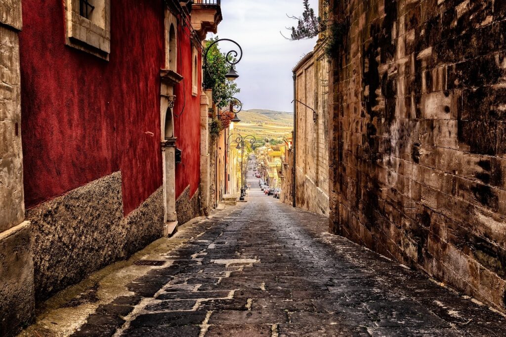 A narrow, cobblestone street in Sicily, Italy, flanked by rustic buildings with red and beige walls, leads down a gentle slope towards a scenic view of the distant countryside. Street lamps and potted plants adorn the sides, enhancing the charming, old-world ambiance.