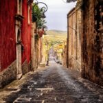 A narrow, cobblestone street in Sicily, Italy, flanked by rustic buildings with red and beige walls, leads down a gentle slope towards a scenic view of the distant countryside. Street lamps and potted plants adorn the sides, enhancing the charming, old-world ambiance.