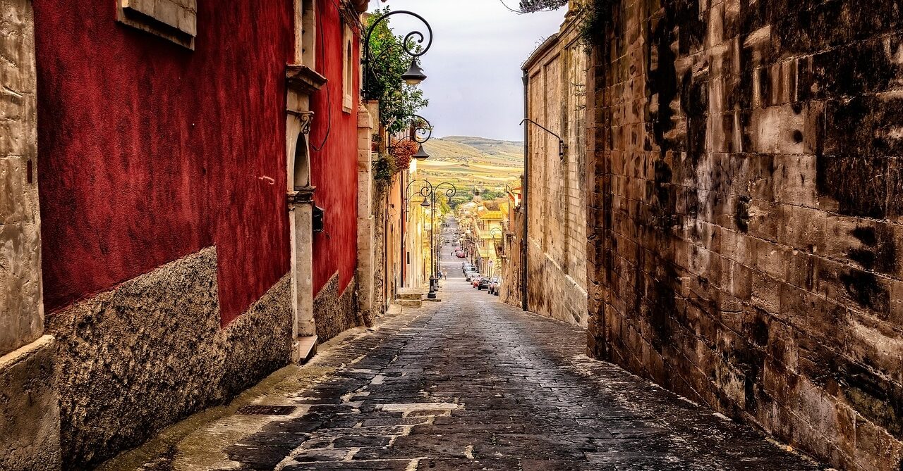 A narrow, cobblestone street in Sicily, Italy, flanked by rustic buildings with red and beige walls, leads down a gentle slope towards a scenic view of the distant countryside. Street lamps and potted plants adorn the sides, enhancing the charming, old-world ambiance.