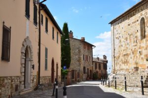 Street view in Cortona, Italy, featuring historic stone buildings with shuttered windows and a large stone wall under a clear blue sky.