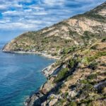 View of the coastline of Elba, featuring a clear blue sea, rocky cliffs, and a small coastal village nestled at the base of rugged hills under a partly cloudy sky