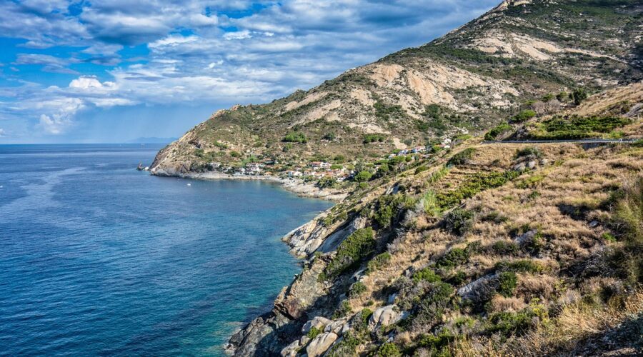 View of the coastline of Elba, featuring a clear blue sea, rocky cliffs, and a small coastal village nestled at the base of rugged hills under a partly cloudy sky