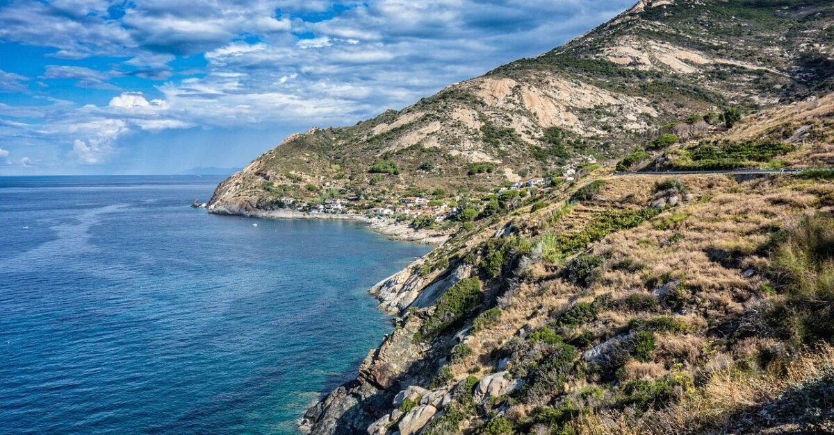 View of the coastline of Elba, featuring a clear blue sea, rocky cliffs, and a small coastal village nestled at the base of rugged hills under a partly cloudy sky