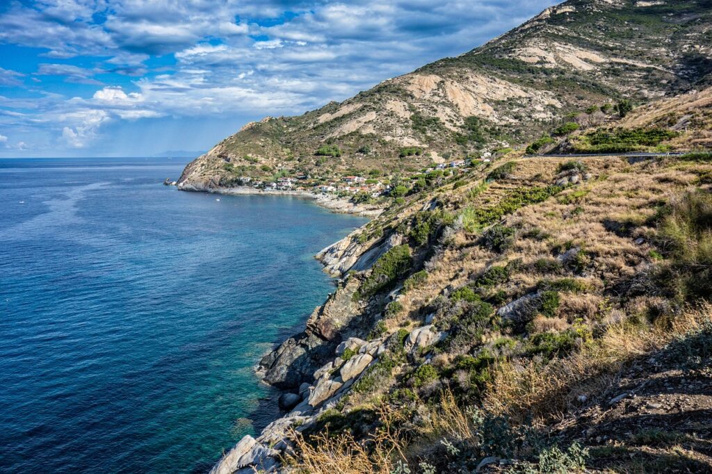 View of the coastline of Elba, featuring a clear blue sea, rocky cliffs, and a small coastal village nestled at the base of rugged hills under a partly cloudy sky.