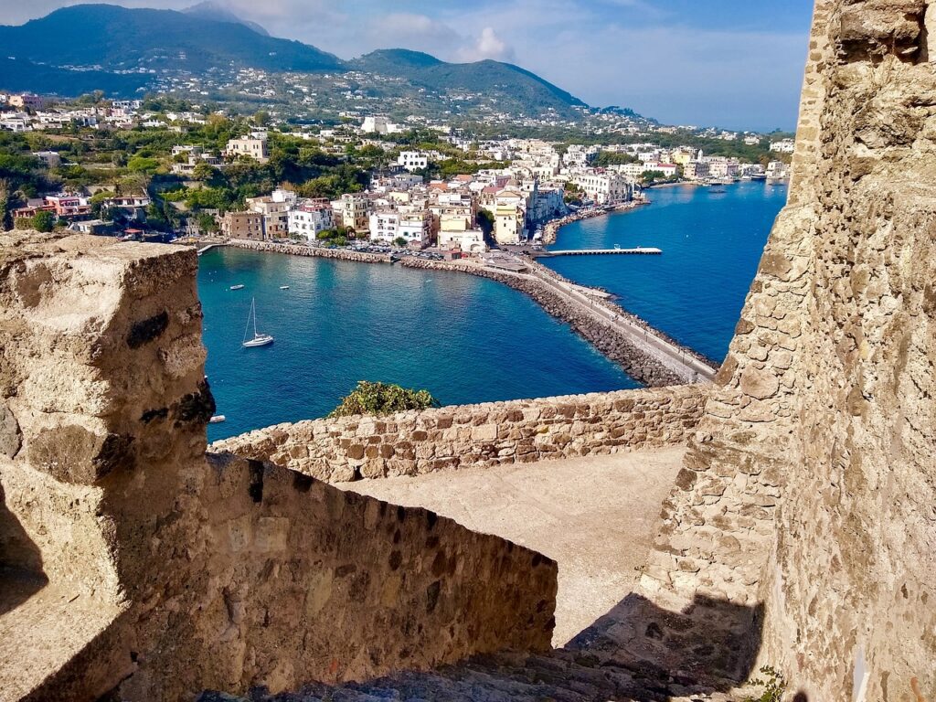 A view of the coastal town of Ischia, one of the Top 5 Italian Islands, with colorful buildings along the shoreline, a calm blue sea, and mountainous terrain in the background, seen from an ancient stone structure. This picturesque setting highlights Ischia's charming architecture and stunning natural landscapes.