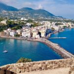 A view of the coastal town of Ischia, with colorful buildings along the shoreline, a calm blue sea, and mountainous terrain in the background, seen from an ancient stone structure. This picturesque setting highlights Ischia's charming architecture and stunning natural landscapes.