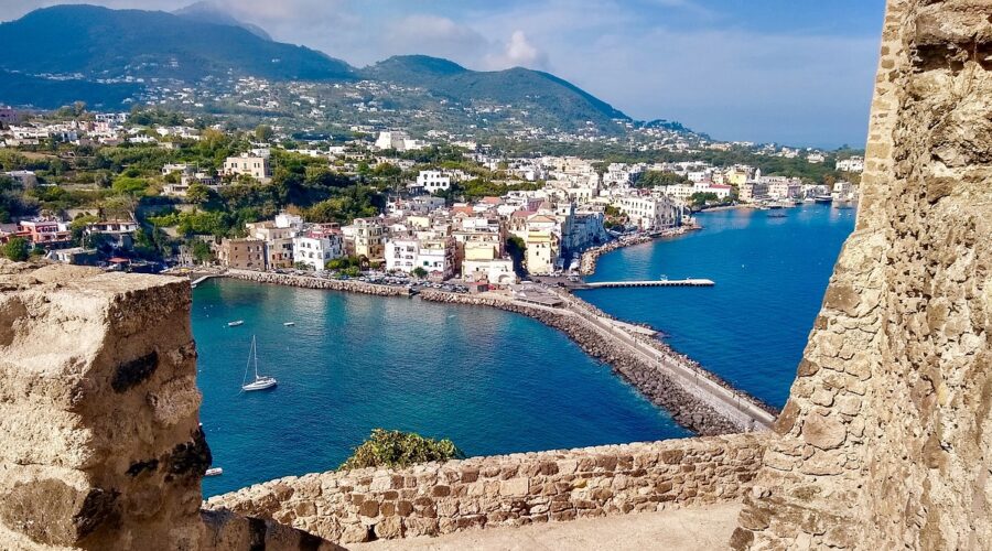 A view of the coastal town of Ischia, with colorful buildings along the shoreline, a calm blue sea, and mountainous terrain in the background, seen from an ancient stone structure. This picturesque setting highlights Ischia's charming architecture and stunning natural landscapes.