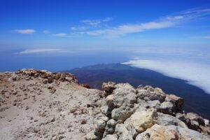 A breathtaking view from the summit of Mount Pico, showing rugged rocks in the foreground and a vast expanse of clouds and ocean below, with a clear blue sky above. (pico Azores travel guide)