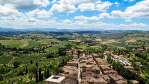 Aerial view of San Gimignano, Italy, showcasing its historic buildings and surrounding lush green vineyards under a blue sky with scattered clouds.