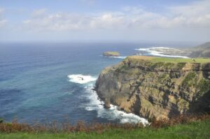 Breathtaking coastal cliffs of São Miguel Island with waves crashing against the rocks and a distant view of a small island under a partly cloudy sky. This dramatic seascape captures the essence of the São Miguel travel guide.