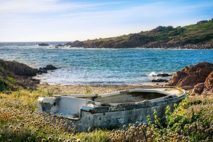 An abandoned boat sits amidst wildflowers on a grassy shore, overlooking the clear blue waters and rocky coastline of Sardinia, one of the Top 5 Italian Islands, under a bright blue sky. This scene captures the island's rugged charm and pristine natural beauty.