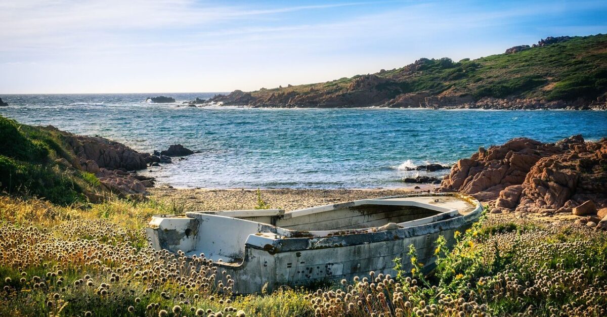 An abandoned boat sits amidst wildflowers on a grassy shore, overlooking the clear blue waters and rocky coastline of Sardinia, one of the Top 5 Italian Islands, under a bright blue sky. This scene captures the island's rugged charm and pristine natural beauty.