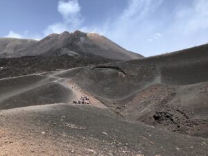 A view of Mount Etna in Sicily, with people walking along the trails on its rugged, volcanic landscape under a clear blue sky.