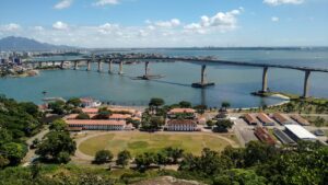 Panoramic view of Terceira Island featuring a long bridge over a calm body of water, with buildings and green areas in the foreground and a distant cityscape and mountains under a blue sky. This image illustrates key attractions highlighted in the Terceira travel guide