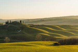 A serene landscape of rolling green hills in Tuscany at sunset, with a distant farmhouse surrounded by cypress trees perched on a hill.