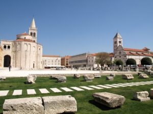 View of a historical square in Zadar, Croatia, with ancient ruins in the foreground, the round Church of St. Donatus, and the Cathedral of St. Anastasia in the background.