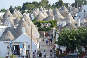 View of the conical-roofed trulli houses in Alberobello, Italy, with people walking through the narrow streets surrounded by these unique stone buildings under a clear sky.