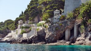 A scenic view of Dubrovnik's coastal architecture featuring stone buildings with ornate gazebos nestled into the cliffside, surrounded by lush greenery, and overlooking clear blue waters.