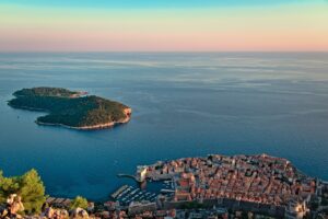 Aerial view of Lokrum Island and the Old Town of Dubrovnik, Croatia, surrounded by the Adriatic Sea under a pastel sunset sky.