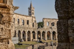 Ancient stone amphitheater with arched windows and a tall bell tower in Pula, Croatia, viewed through a gap in the ruins.