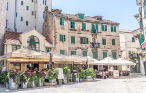 Outdoor seating area of Tavern Faros in Split, Croatia, with patrons dining under umbrellas and a charming old building with green shutters in the background.