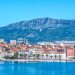 Panoramic view of Split, Croatia, with its colorful buildings and red-tiled roofs along the waterfront, set against a backdrop of towering mountains under a clear blue sky.