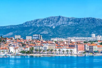 Panoramic view of Split, Croatia, with its colorful buildings and red-tiled roofs along the waterfront, set against a backdrop of towering mountains under a clear blue sky.