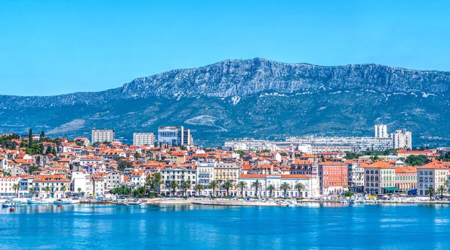 Panoramic view of Split, Croatia, with its colorful buildings and red-tiled roofs along the waterfront, set against a backdrop of towering mountains under a clear blue sky.