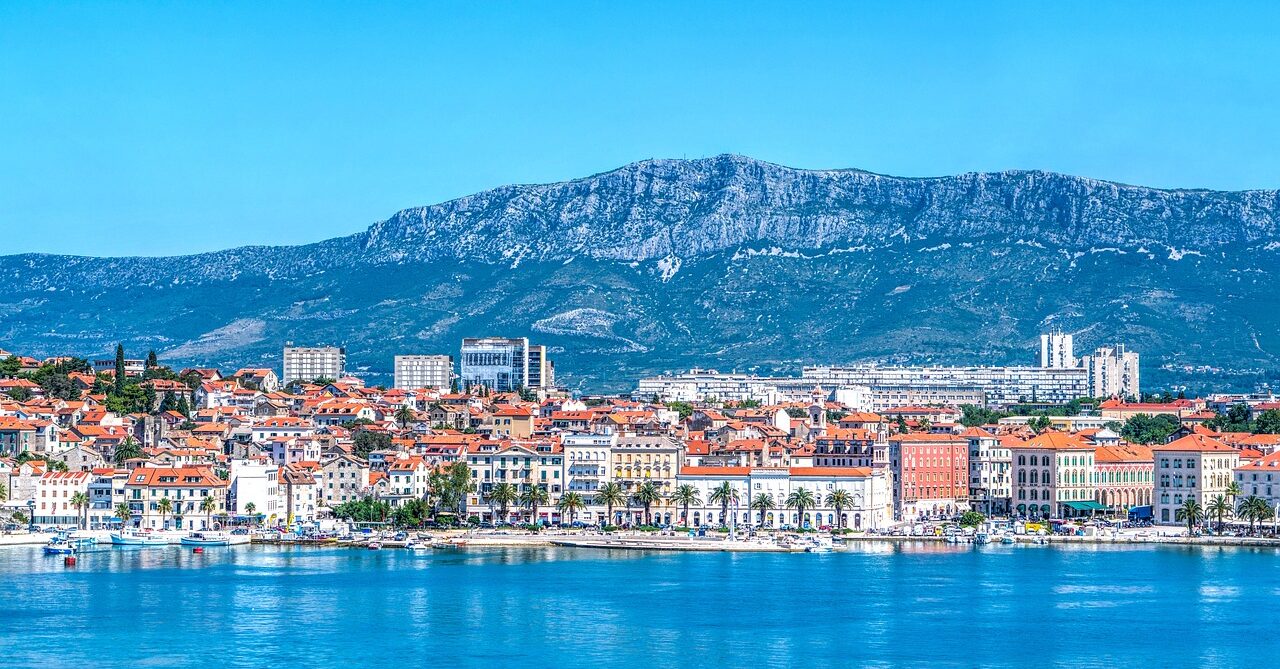 Panoramic view of Split, Croatia, with its colorful buildings and red-tiled roofs along the waterfront, set against a backdrop of towering mountains under a clear blue sky.