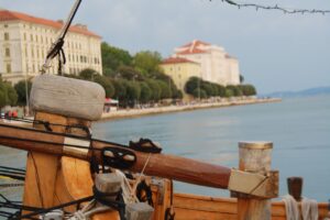 Wooden mast and rigging of a traditional ship with a scenic view of Zadar's waterfront, featuring historic buildings and lush greenery along the promenade.