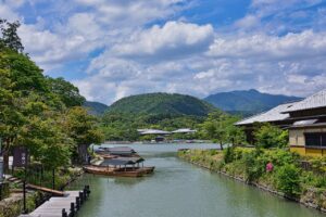 Scenic view of a river with traditional boats docked along the shore, surrounded by lush greenery and mountains under a partly cloudy sky in Arashiyama, Kyoto, Japan.