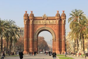 A view of the Arc de Triomf in Barcelona, Spain, a large brick archway with intricate carvings, surrounded by palm trees and people walking along the promenade.