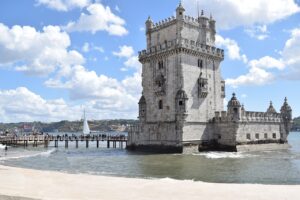 The historic Belém Tower in Lisbon, Portugal, standing against a bright blue sky with fluffy clouds, with a crowd of visitors on the bridge and a sailboat in the background.