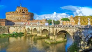 View of Castel Sant'Angelo and the Sant'Angelo Bridge in Rome, with clear blue skies and reflections in the Tiber River.