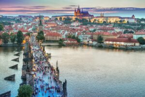 Aerial view of the Charles Bridge in Prague, crowded with tourists, leading to the illuminated Prague Castle at sunset.