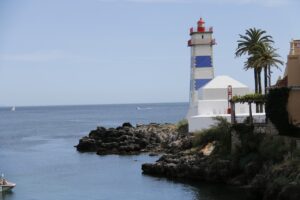 A striped lighthouse stands by the rocky shore, with palm trees and the calm ocean stretching into the horizon, and a small boat floating nearby.