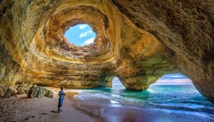 A person stands inside the stunning Benagil Cave in Algarve, Portugal, with its large natural skylight and view of the ocean through the cave's arches.