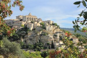 Scenic view of the historic hilltop village of Gordes in Provence, France, with traditional stone buildings cascading down the hillside, surrounded by lush greenery and framed by red berries in the foreground.