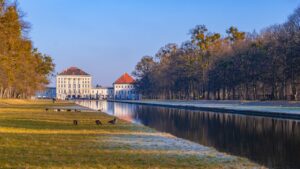 View of Nymphenburg Palace in Munich, Germany, with its reflective canal, surrounding greenery, and grazing geese on a clear autumn day.