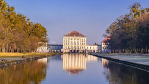 The Nymphenburg Palace in Munich reflected in the calm waters of its canal, surrounded by lush trees under a clear blue sky.