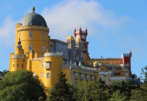 Colorful and intricate Pena Palace in Sintra, Portugal, with its yellow, red, and gray towers and domes, set against a blue sky and surrounded by lush greenery.